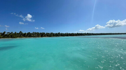 Ocean or sea calm water surface under a blue sky with a few clouds. View on the shore with a beach and tropical trees. Vacation resort paradise in summer time