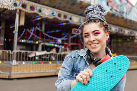 lifestyle portrait extraordinary hipster woman with african braids dressed in a denim jacket with a skateboard in hand at amusement park