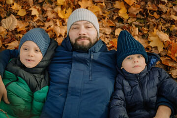 happy father and his two sons lying on the ground covered with yellow fallen leaves in a park on sunny autumn day 