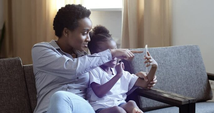 Happy family african american ama nanny black woman older sister sitting with cute baby girl daughter on couch in living room speaks into web camera of phone looking at smartphone screen make photo