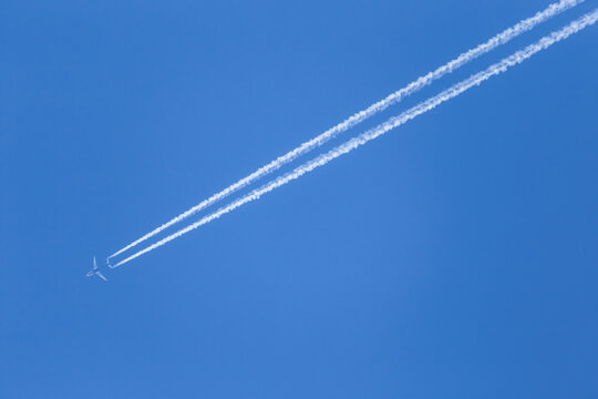 Airplane is flying high in the clear blue sky. Two white condensation trail cross air space.