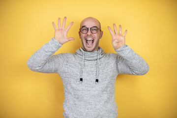 Young bald man wearing glasses over yellow background showing and pointing up with fingers number...