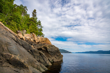 Fototapeta na wymiar Huge yellow rocks with green trees flowing down the sea of Fjord du Saguenay in Quebec, Canada, during a cloudy day