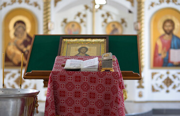 Closeup interior of a Christian church. Prayer table covered with patterned red cloth. A metal container with holy water for baptism. Bible, casket, icon and blurred iconostasis in the background.
