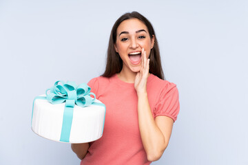 Pastry chef holding a big cake isolated on blue background shouting with mouth wide open