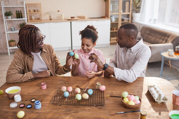 Fototapeta premium High angle portrait of happy African-American family painting Easter eggs together while sitting at wooden table in cozy home interior, DIY Easter decorations