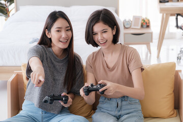Two Asian girlfriends enjoy playing video games with joysticks at home. Lesbian couples or lgbt activities on vacation inside the room.