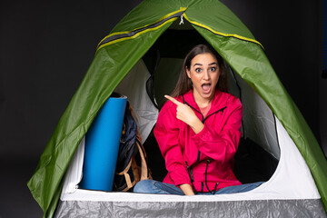 Young caucasian woman inside a camping green tent surprised and pointing side