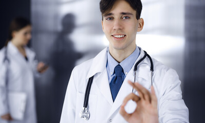 Friendly man-doctor standing straight and showing Ok sign in clinic. Female colleague is in a hurry at the background of physician. Medicine concept