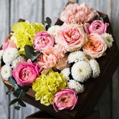Wooden basket in the shape of a heart with pink roses and white, yellow flowers stands on a stand against a gray background