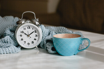 Gray knitted plaid, blue mug and alarm clock on a white background. Seasonal time change, daylight saving time concept.
