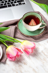  A cup of tea, tulips and a cake next to a laptop on the table. Cozy work from home concept in spring, vertical photo