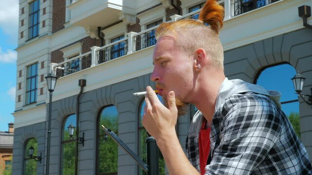 A hipster smoking a cigarette outdoors. A young man smoking in the city near a modern building or a cultural center. Bad habits and youth culture concept