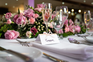 Groom wedding breakfast place setting with name card on top table waiting for arrival with bridge and pink roses and shiny champagne glasses