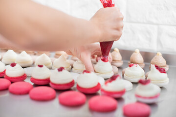 Obraz na płótnie Canvas Close up view of female hands of professional pastry chef, holding confectionery bag and squeezing red fruit jam on white macarons shells with buttercream