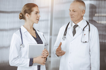 Elderly confident doctor and woman-physician discussing current disease therapy while standing in sunny clinic. Teamwork in medicine