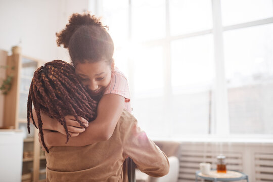 Minimal waist up portrait of happy African-American girl embracing mother in cozy home interior lit by sunlight, copy space