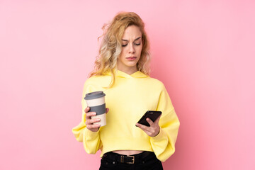 Young blonde woman wearing a sweatshirt isolated on pink background holding coffee to take away and a mobile