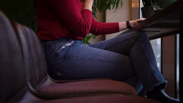 Woman in blue jeans red sweater sits high bar stool, cross-legged table downtown coffee shop. Drinks tea from large green cup. Enjoy cozy time spent, moment, a day off, a lunch break. Below the belt