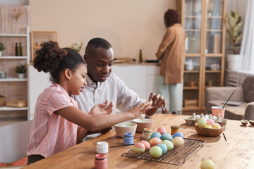 Portrait of young African-American girl painting Easter eggs while enjoying DIY decorating with loving father, copy space