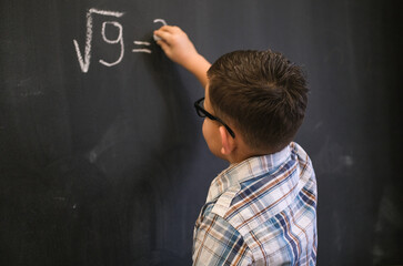 cute schoolboy boy solves a math problem on a black blackboard. calculating the square root