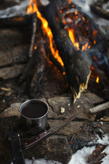 Metal mug with tea and a knife lying on a stone on the background of a burning fire , close-up.