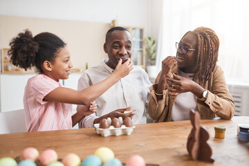 Portrait of loving African-American family making chocolate Easter decorations and tasting them while sitting at wooden table in cozy home interior