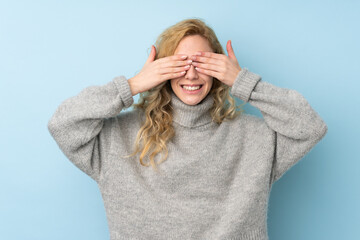 Young blonde woman wearing a sweater isolated on blue background covering eyes by hands