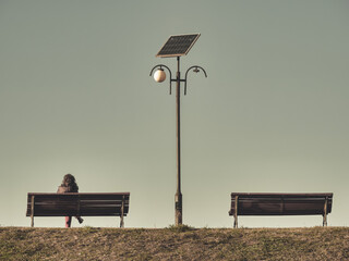 Lonely woman sitting on a bench next to a light pole. Two benches with a solar panel lamp post in...