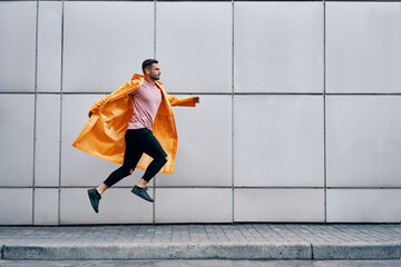 Trendy young man in yellow raincoat jumping in the air on wall street urban background