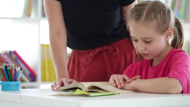 Little girl reading book under teacher supervision sitting at school desk