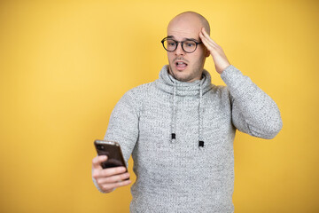 Young bald man wearing glasses over yellow background chatting, surprised and looking his phone