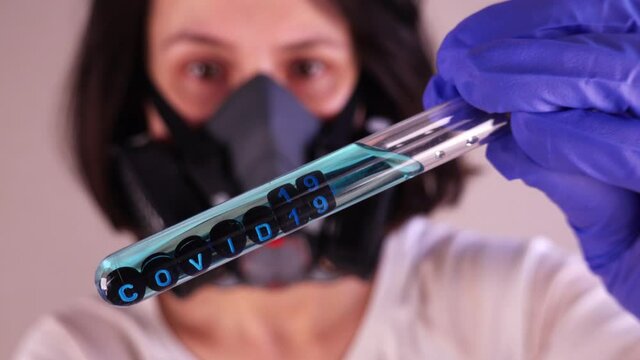 Caucasian Female Scientist Wearing Gas Mask and Blue Latex Gloves while Examining Test Tube With Covid-19 Sample
