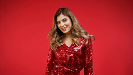 Fototapeta premium positive woman in shiny dress looking at camera isolated on red