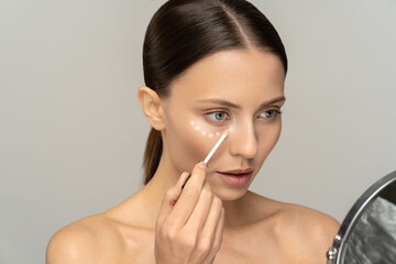 Woman with natural makeup applying concealer on flawless fresh skin, doing make up, looking at...