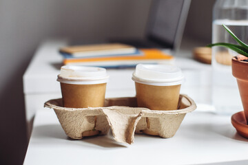 Two paper take away coffee cups in cardboard holder. Laptop, notebooks, plant, water on a white table