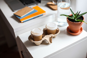 Two paper take away coffee cups in cardboard holder. Laptop, notebooks, plant, water on a white table
