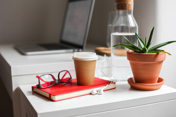 Paper take away coffee cup, red notebook, glasses, white earphones, aloe plant, a jar of water on a white table, workspace 