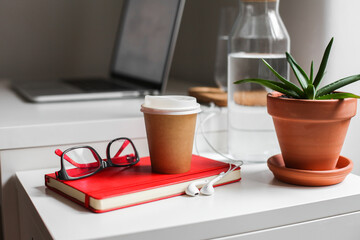 Paper take away coffee cup, red notebook, glasses, white earphones, aloe plant, a jar of water on a white table, workspace 