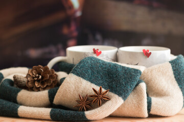 Romantic Love Nature Mood Creative Composition. Close-up Warm Green Beige Scarf Tied Two Mugs with Red Hearts. Spicy Drink. Background is firewood in fireplace. Stay warm together.