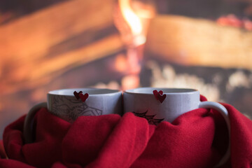 Real Love and Valentine's Day Mood - romantic concept. Close-up Two mugs with double red hearts are tied with one red scarf. Background is hot firewood in fireplace. Creative Still life Composition.