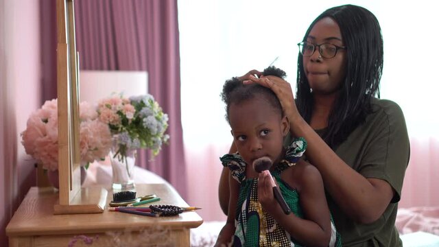 african american mother combing  her little daughter hair sitting near mirror on dressing table in the children room at home. black mom and girl at bedroom in morning time together