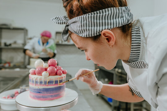 Female Confectioner Decorating Birthday Cake Using Edible Golden Dye And Brush