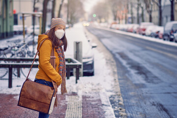 Midde-aged brunette woman standing at a bus stop, wearing a protective face mask due to corona virus, waiting for her bus to bring her to work