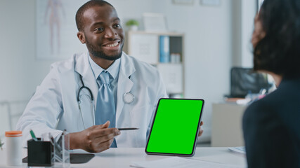 African American Medical Doctor is Explaining Diagnosis to a Patient on a Tablet with Green Screen in a Health Clinic. Assistant in White Lab Coat is Reading Medical History in Hospital Office. 