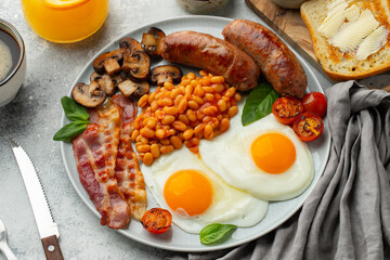 Full English breakfast on a plate with fried eggs, sausages, bacon, beans, toasts and coffee on light stone background. Top view