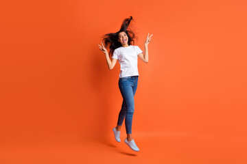 Fototapeta na wymiar Full size photo of optimistic girl jump show v-sign wear white t-shirt jeans sneakers isolated on orange color background