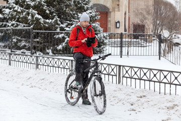 Bicyclist typing screen of smartphone while standing with bike on snowy urban streets, riding on bike at winter season