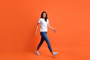 Full size profile photo of optimistic girl go wear white t-shirt jeans sneakers isolated on orange color background