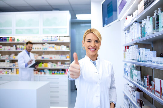Portrait of beautiful female blonde pharmacist holding thumbs up in pharmacy shop or drugstore. In background shelves with medicines. - Powered by Adobe